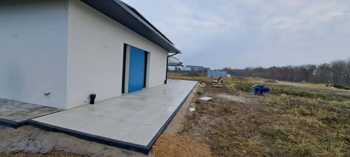 Wyłożony jasnymi płytami taras przy nowoczesnym domu z niebieskimi drzwiami w Rudzie Śląskiej, widok na niezagospodarowany teren z resztkami gruzu i sprzętem budowlanym w tle.