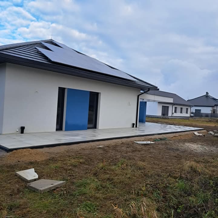 Nowoczesny dom jednorodzinny z panelami słonecznymi na dachu i świeżo ułożonym tarasem z jasnych płyt betonowych, widoczne niedokończone prace ziemne wokół tarasu.