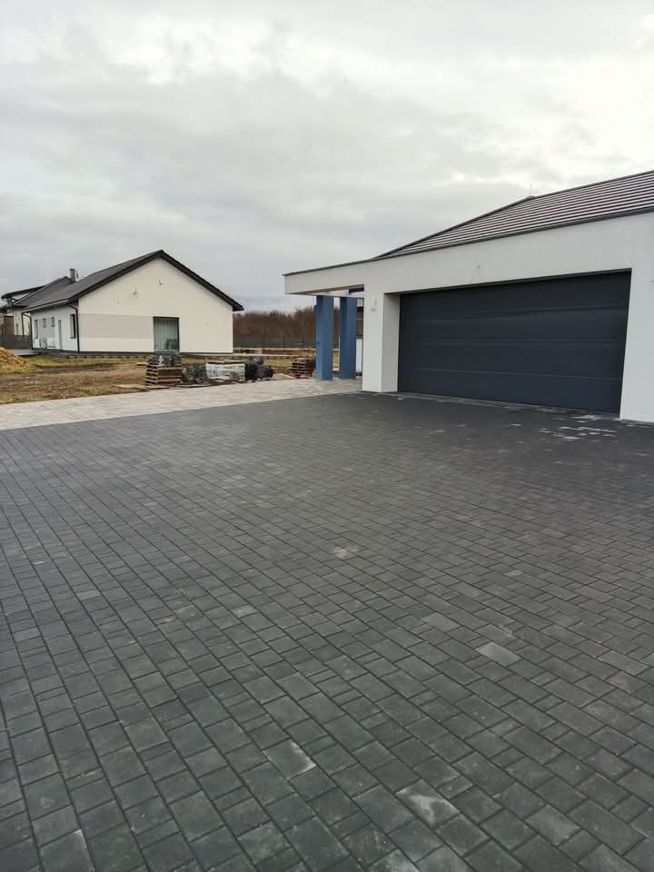 Ułożona, szara kostka brukowa na podjeździe do garażu nowoczesnego domu jednorodzinnego w pochmurny dzień. Widoczny fragment drugiego domu i przygotowane materiały budowlane.