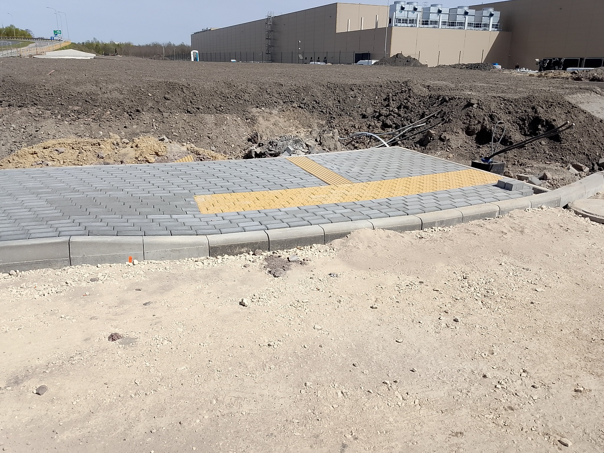 Nowo ułożona szara kostka brukowa z żółtymi pasami dla osób niedowidzących, obrzeża betonowe, teren budowy z ziemią i budynkiem w tle.