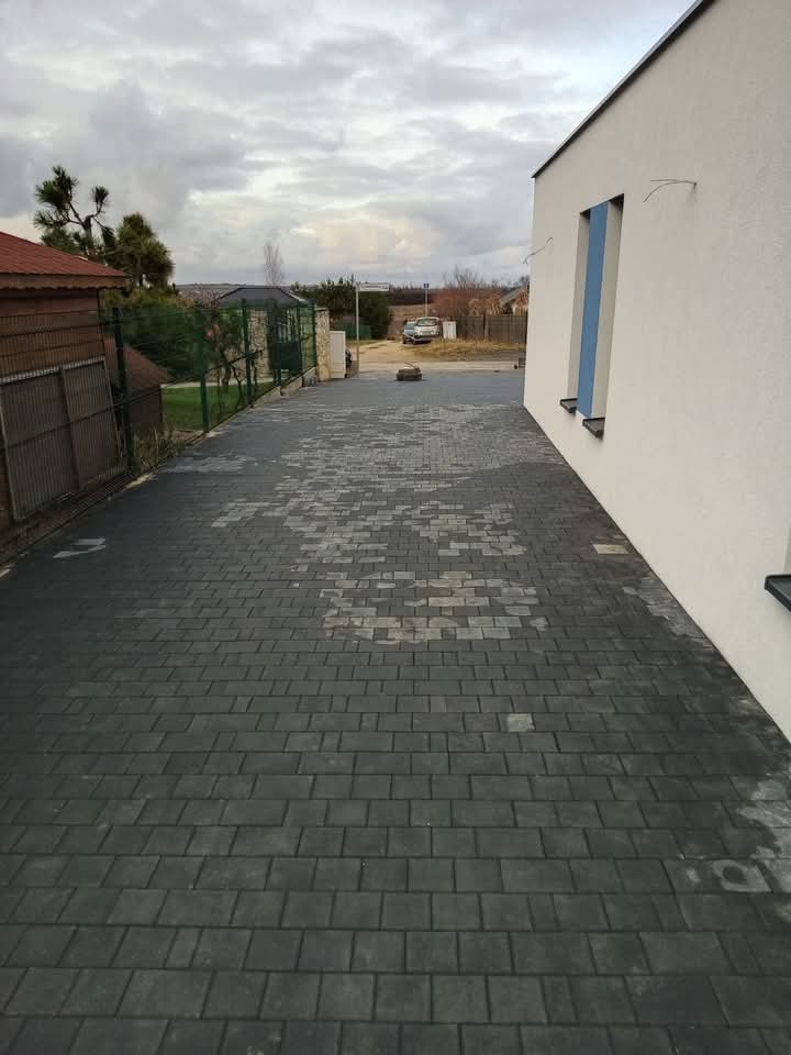 Ułożona, ciemnoszara kostka brukowa na podjeździe, widoczne jaśniejsze przebarwienia i ślady po wodzie, obok biały budynek z niebieskimi wnękami okiennymi, w tle brama wjazdowa i samochód.