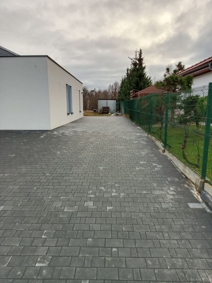 Szeroki podjazd z szarej kostki brukowej ułożonej we wzór jodełki, obok nowoczesny biały dom i zielone ogrodzenie panelowe.