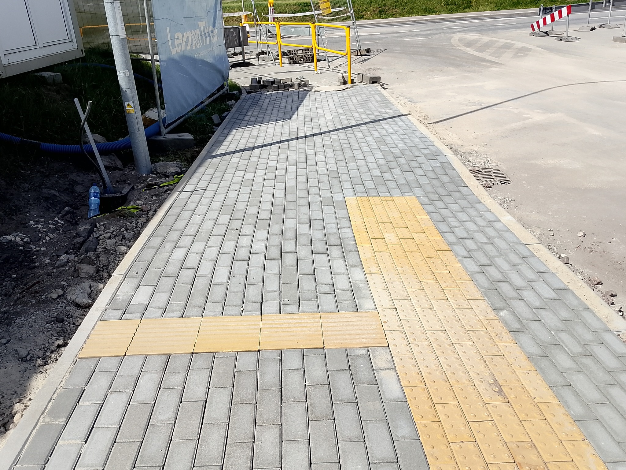 Nowo ułożony chodnik z szarej kostki brukowej z żółtymi pasami dla osób niedowidzących, widoczne krawężniki i elementy infrastruktury drogowej w tle.