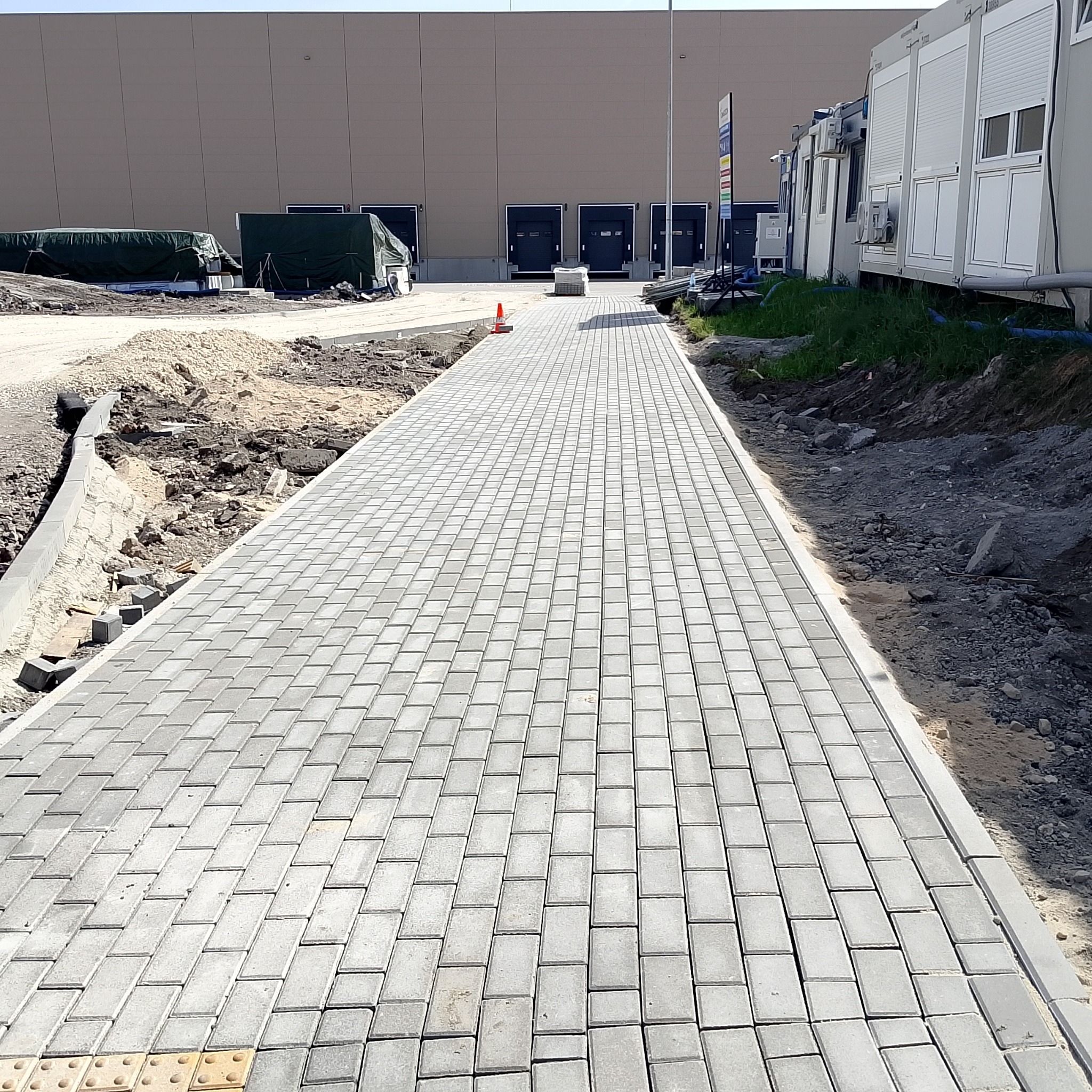 Świeżo ułożona, długa ścieżka z szarej kostki brukowej prowadząca w kierunku magazynu z rampami, widoczne prace wykończeniowe po bokach, pomarańczowy pachołek ostrzegawczy na ścieżce.