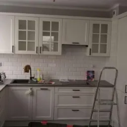 Jasna kuchnia z białymi szafkami z przeszklonymi górnymi frontami i czarnymi uchwytami, biała płytka na ścianie, srebrna drabina oparta o szafki, widok na okno.