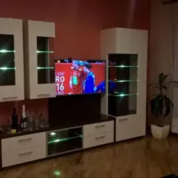 Zestaw mebli salonowych z podświetlanymi półkami, telewizorem i dekoracjami, w tle zasłonięte okno i roślina doniczkowa.