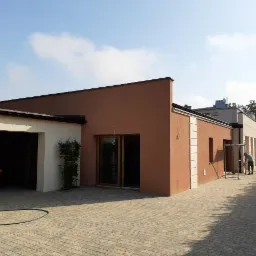 Nowoczesny dom jednorodzinny z garażem i świeżo pomalowaną elewacją w kolorze terakoty, widoczne detale wykończeniowe i ekipa budowlana przygotowująca się do dalszych prac.