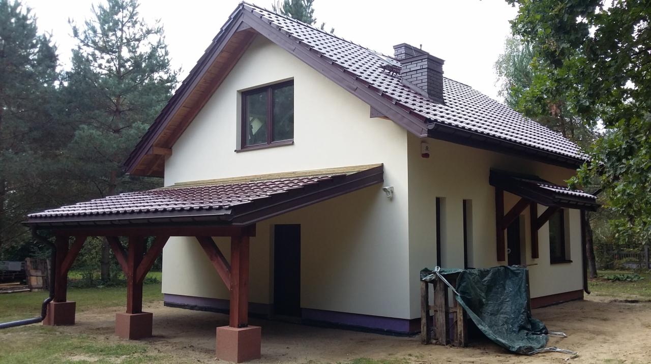 Jasny, dwukondygnacyjny dom z brązowym dachem z dachówki i drewnianą wiatą, świeżo po pracach wykończeniowych, z widocznym fragmentem elewacji i obróbki blacharskiej.