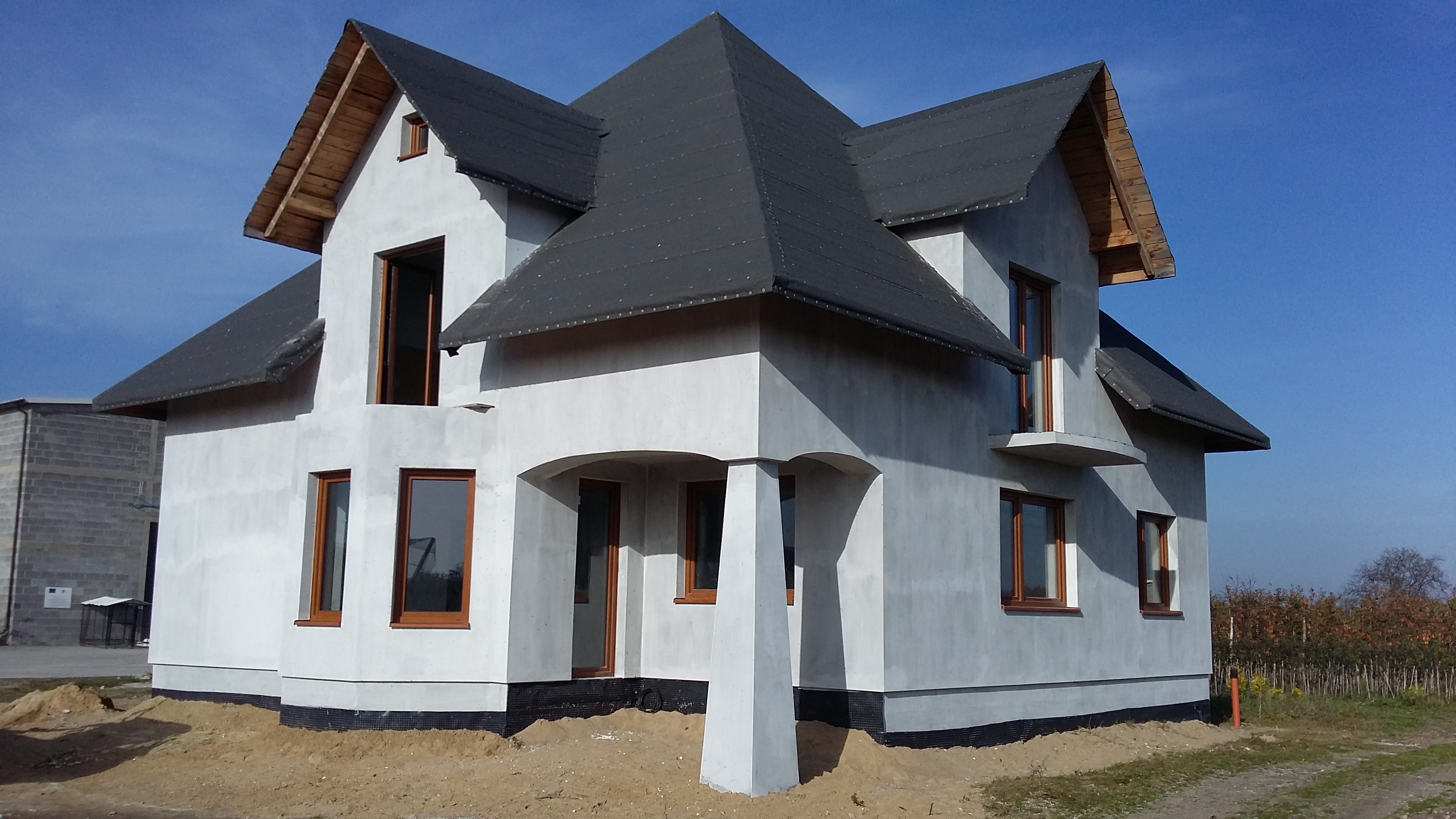 Surowy stan nowo wybudowanego domu z szarą elewacją, ciemnym dachem i drewnianymi ramami okiennymi, widoczne niedokończone otoczenie.