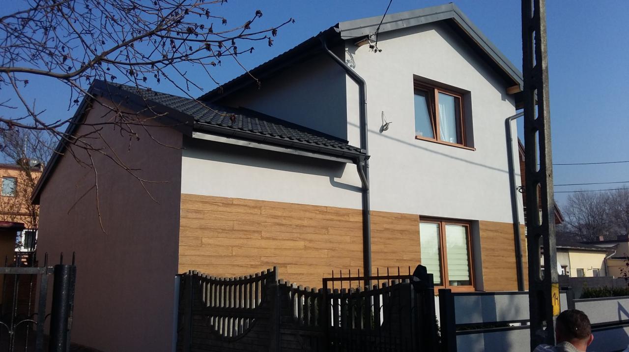 Dwukondygnacyjny dom jednorodzinny z jasnoszarą elewacją górnej części i okładziną imitującą drewno w dolnej części, widoczny zza ogrodzenia w słoneczny dzień.