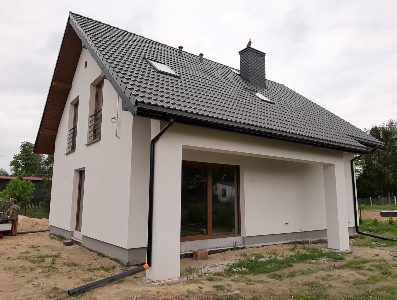 Nowo wykończony dom jednorodzinny z jasną elewacją, ciemną dachówką i elementami wykończeniowymi w odcieniach brązu, widoczny fragment terenu wokół domu z elementami trawy i piasku.