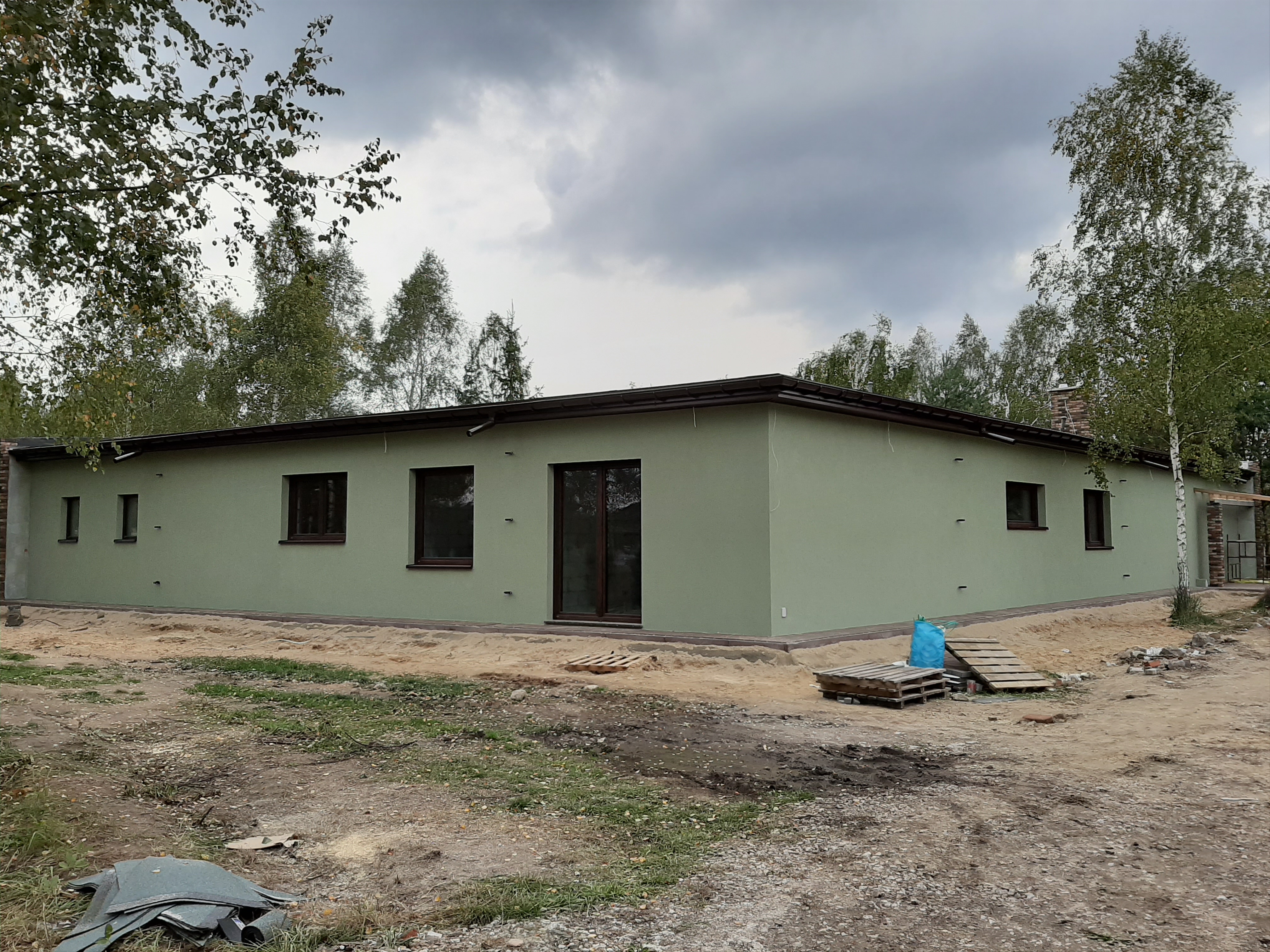 Jednopiętrowy dom z zieloną elewacją i brązowymi ramami okiennymi, otoczony drzewami i placem budowy z paletami i workiem na śmieci.