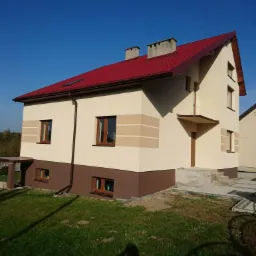 Nowo wybudowany, dwukondygnacyjny dom z czerwoną dachówką, beżową elewacją z brązowym pasem u dołu i jasnymi paskami dekoracyjnymi, widoczny z perspektywy trawnika w słoneczny dzień.