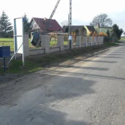 Nowoczesne ogrodzenie z szarej kostki łupanej z regularnymi słupkami, widoczne skrzynki pocztowe i tablica informacyjna przy drodze w wiejskim krajobrazie.