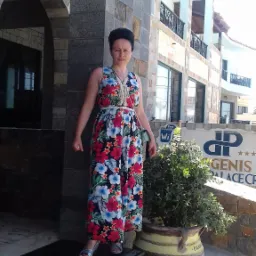 Kobieta w kwiecistej sukience stoi na schodach przed wejściem do hotelu Genis Free Palace Crete, obok donicy z krzewem.