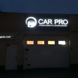 Elewacja budynku z podświetlanym szyldem 'CAR PRO Studio Kosmetyki Samochodowej' nad bramą garażową i mniejszym szyldem 'Myjnia LUX' obok drzwi wejściowych.