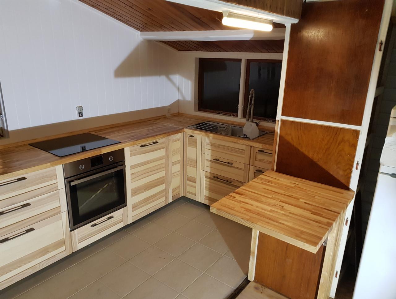 Jasna kuchnia z drewnianymi frontami szafek o wyraźnym usłojeniu, czarny piekarnik i płyta indukcyjna w zabudowie, blat z drewna, rozkładany stolik przymocowany do boku szafki, szara podłoga z płytek.