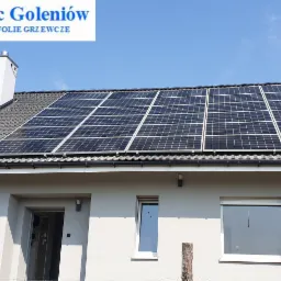 Panele fotowoltaiczne zamontowane na dachu domu jednorodzinnego z szarą dachówką, widoczne rynny i komin. Na górze logo firmy.