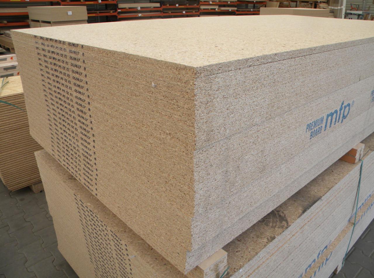 Stos płyt drewnopochodnych z widocznym napisem 'PREMIUM BOARD mfp' ułożonych w magazynie, widoczne krawędzie i struktura materiału, w tle regały z innymi materiałami budowlanymi.