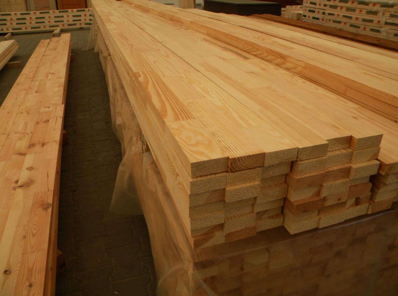 Stos belek z jasnego drewna ułożonych na palecie, widoczne słoje i struktura drewna, w tle inne belki i palety.
