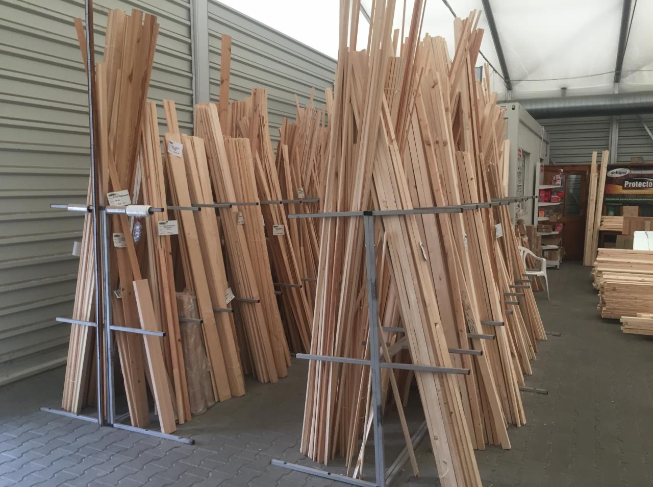 Stosy jasnego drewna budowlanego ułożone w metalowych regałach w magazynie, widoczne etykiety z cenami i wymiarami.