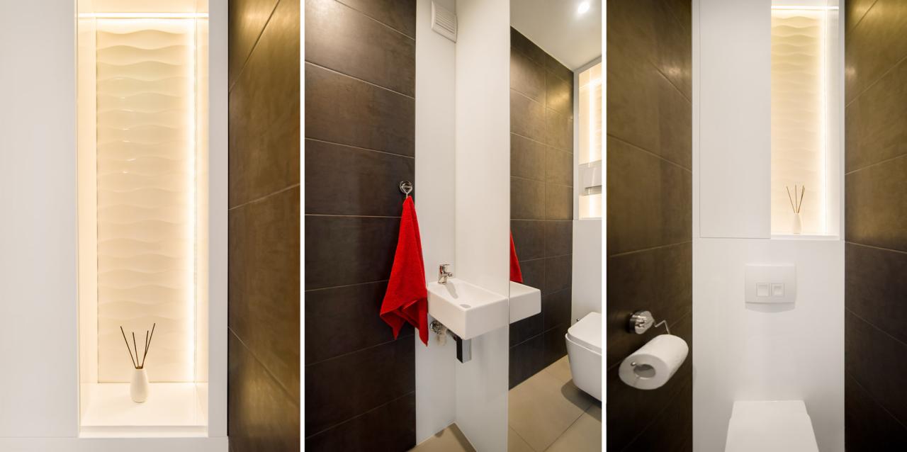 Nowoczesna łazienka z ciemnymi płytkami, podświetloną wnęką z dekoracyjną ścianą i dyfuzorem zapachowym, białą umywalką z czerwonym ręcznikiem oraz podwieszaną toaletą.