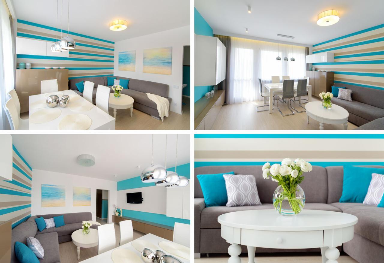 Wnętrze salonu z aneksem jadalnym w odcieniach bieli, szarości i turkusu, z poziomymi pasami na ścianach, szarym narożnikiem z turkusowymi poduszkami, białym stolikiem kawowym z bukietem białych...