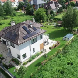 Widok z góry na nowoczesny dom jednorodzinny z brązowym dachem i panelami fotowoltaicznymi, otoczony zielonym ogrodem z oczkiem wodnym i altaną, na podjeździe stoi ciemny samochód.