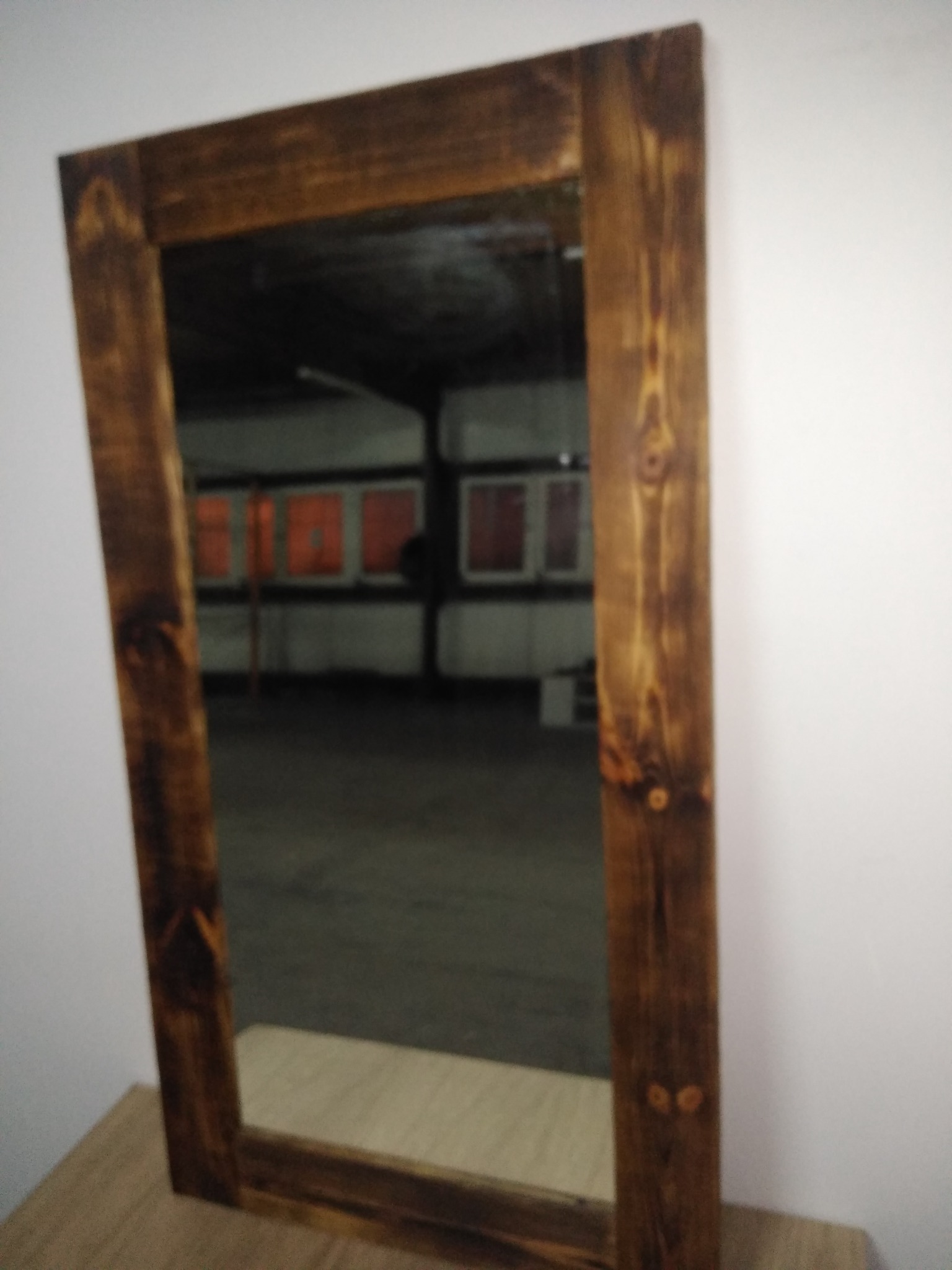 Drewniane lustro w prostej, ciemnej ramie z widocznymi sękami, ustawione na blacie, odbijające wnętrze warsztatu z czerwonymi oknami.