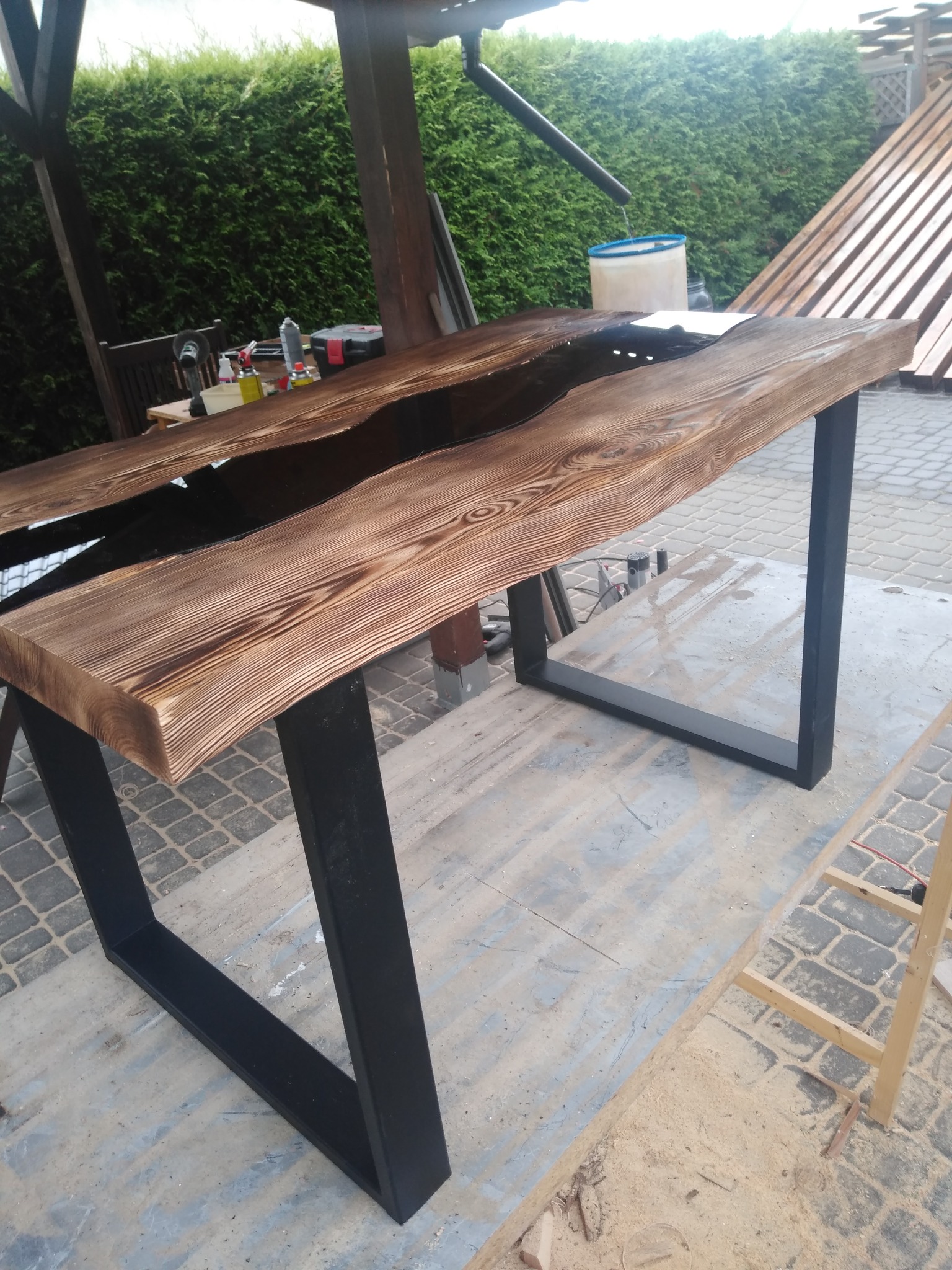 Brązowy blat drewniany z żywicą epoksydową w kolorze czarnym, osadzony na czarnych, metalowych nogach w kształcie litery U, widoczny w warsztacie z narzędziami i zielenią w tle.