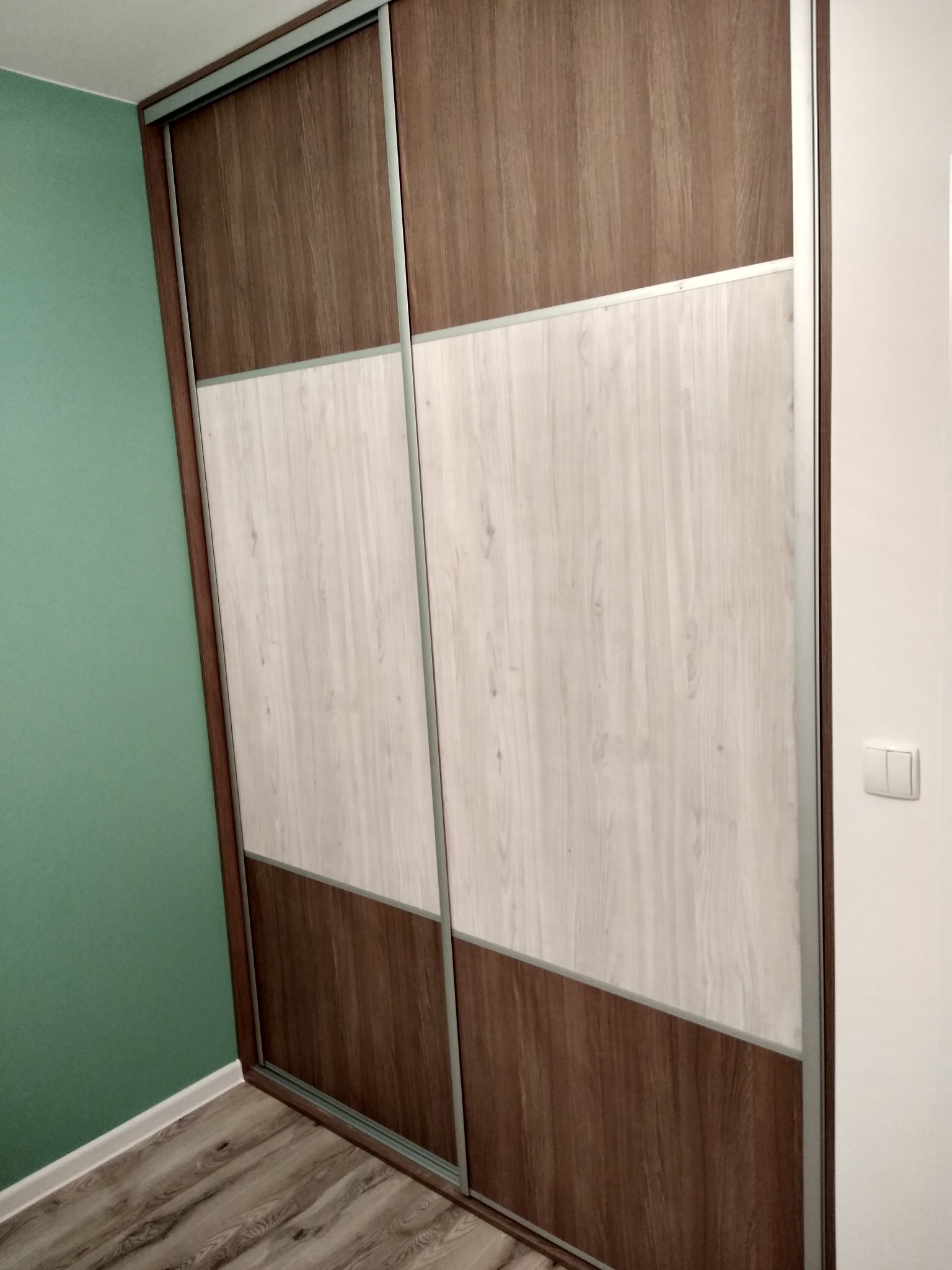 Dwudrzwiowa szafa wnękowa z przesuwnymi drzwiami, łącząca panele w kolorze jasnego i ciemnego drewna, umieszczona w rogu pokoju z zieloną ścianą i drewnianą podłogą.