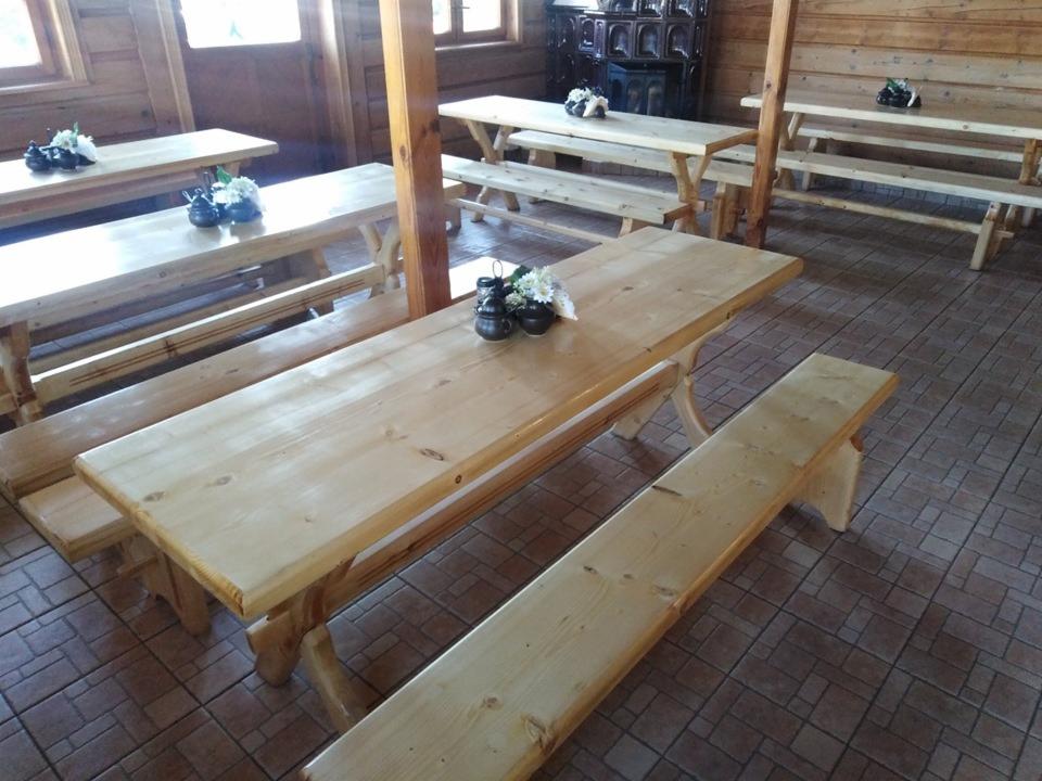 Wnętrze drewnianej sali z kilkoma stołami i ławami wykonanymi z jasnego drewna, na stołach dekoracje z kwiatów, w tle piec kaflowy.