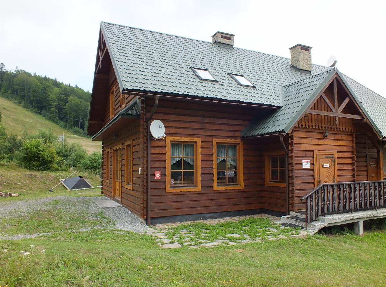 Drewniany budynek z brązowych bali z szarym dachem z blachy trapezowej, widoczne okna z drewnianymi ramami, kominy, antena satelitarna oraz fragment zbocza góry z zielonym lasem w tle.