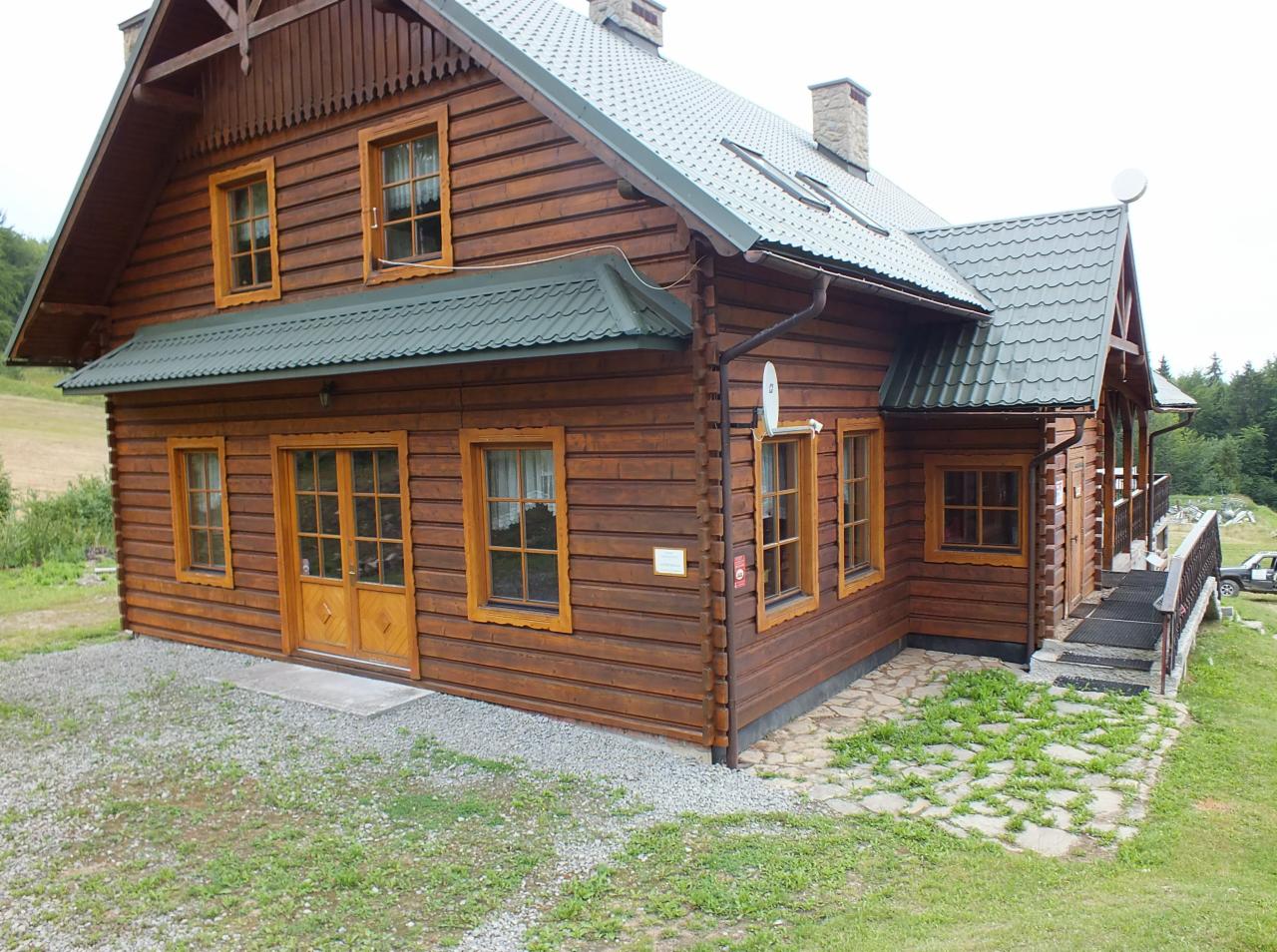 Gotowy, dwukondygnacyjny dom z bali drewnianych z zielonym dachem, widoczny z boku, z fragmentem trawnika i ścieżki wyłożonej kamieniami.