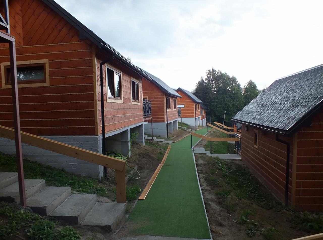 Rząd drewnianych domków letniskowych na podwyższeniu, połączonych ścieżką z zielonej sztucznej trawy, betonowe schody prowadzące do jednego z domków.
