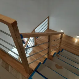 Widok z góry na zabezpieczone schody z drewnianymi poręczami i metalowymi prętami, jasna drewniana podłoga i białe ściany z punktowym oświetleniem.