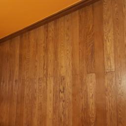 Stolarstwo Marek Wiśniewski - Ułożona pod kątem podłoga z desek o ciepłym, brązowym odcieniu, widoczna z perspektywy pomieszczenia z pomarańczowym sufitem i fragmentem ciemnobrązowych drzwi.