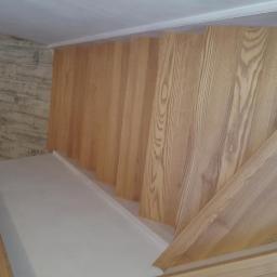 Stolarstwo Marek Wiśniewski - Widok z góry na drewniane schody z jasnego drewna z widocznym butem na jednym ze stopni, ściana wyłożona imitacją drewnianych paneli.