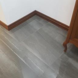 Stolarstwo Marek Wiśniewski - Szare płytki podłogowe z drewnianą listwą przypodłogową i fragmentem drewnianego mebla w rogu.