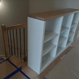 Stolarstwo Marek Wiśniewski - Biały regał z drewnianym blatem umieszczony na podeście schodów z drewnianą balustradą z czarnymi prętami, podłoga zabezpieczona kartonem i taśmą malarską, widoczne przygotowania do prac...