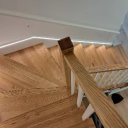 Drewniane schody ze skrętem, oświetleniem LED pod stopniami, białą balustradą i drewnianą poręczą, widziane z góry, z lekkim bałaganem na podłodze obok poręczy.