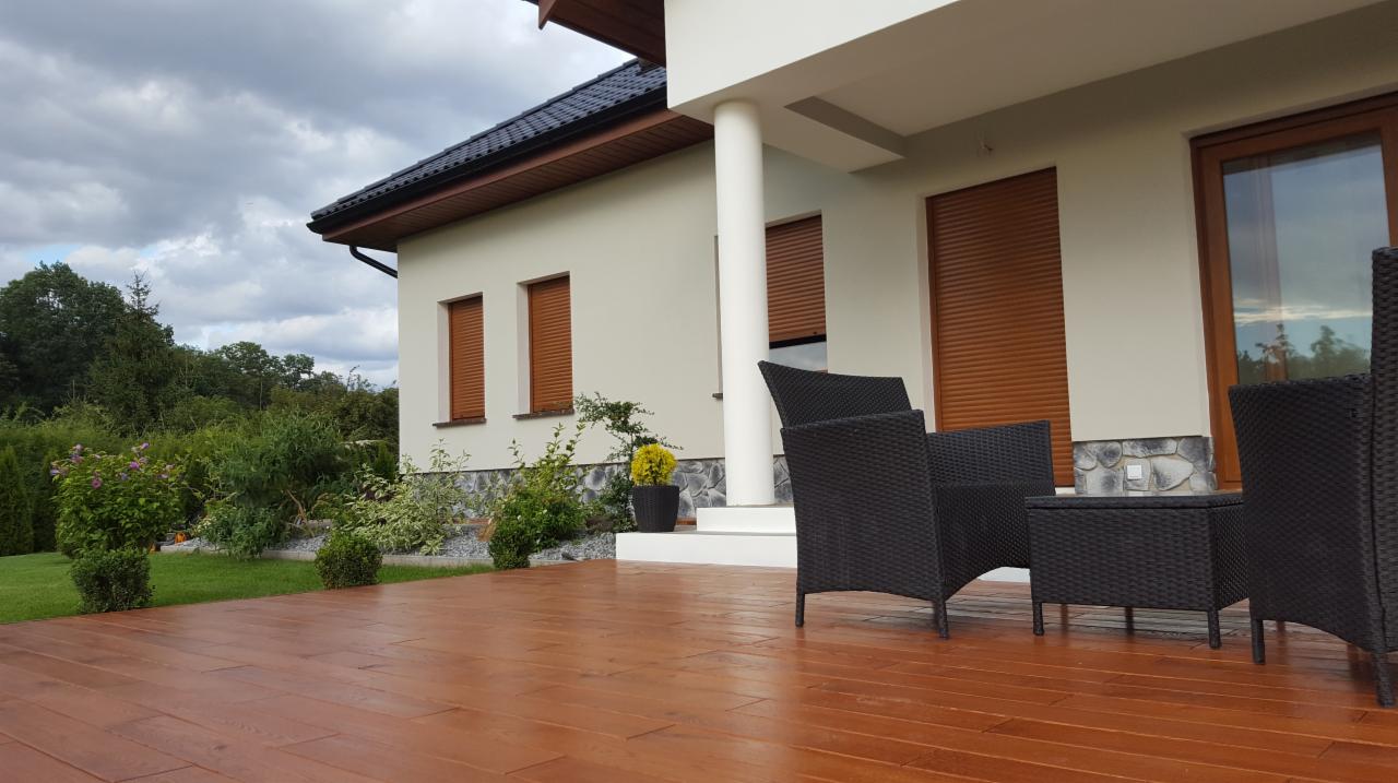 Brązowy taras z deskami kompozytowymi, meble ogrodowe z rattanu w kolorze czarnym, kremowa elewacja domu z brązowymi roletami zewnętrznymi i kolumną przy wejściu, zadbany trawnik i ogród w tle.