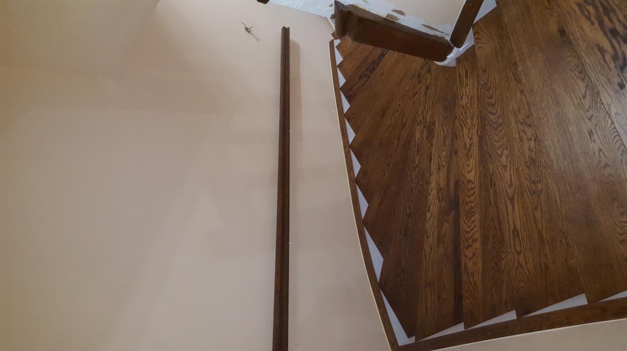 Wnętrze domu w trakcie montażu ciemnobrązowych schodów z drewna, widoczne przygotowane stopnie i listwa przypodłogowa, ściana w odcieniu beżu.