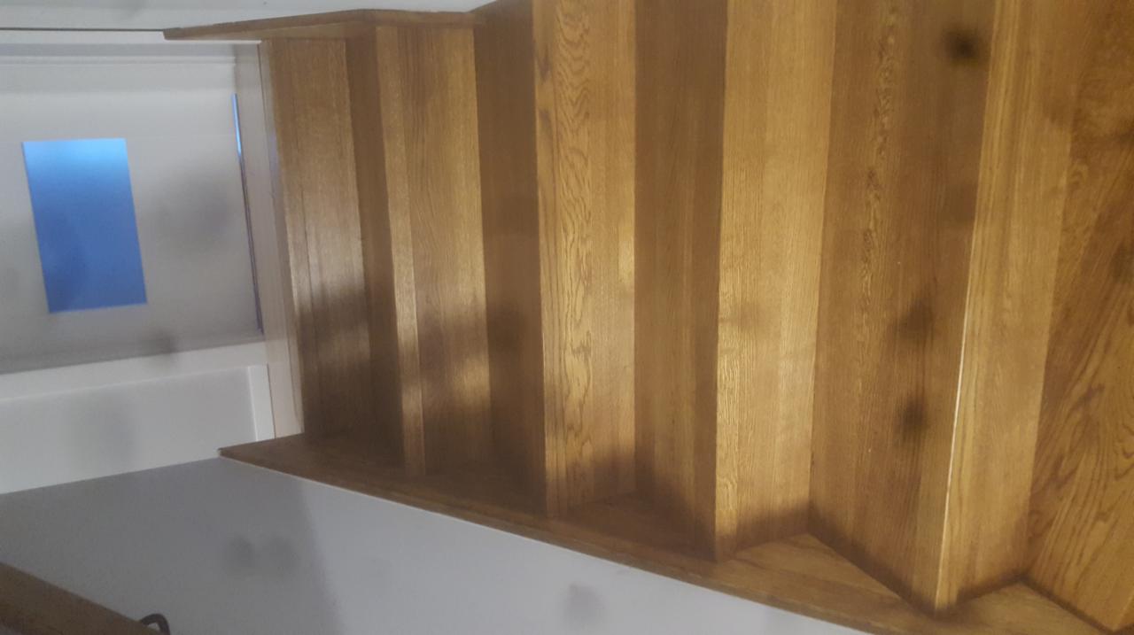 Fragment konstrukcji schodów z jasnego drewna, widoczne pionowe elementy i fragment ściany z oknem.