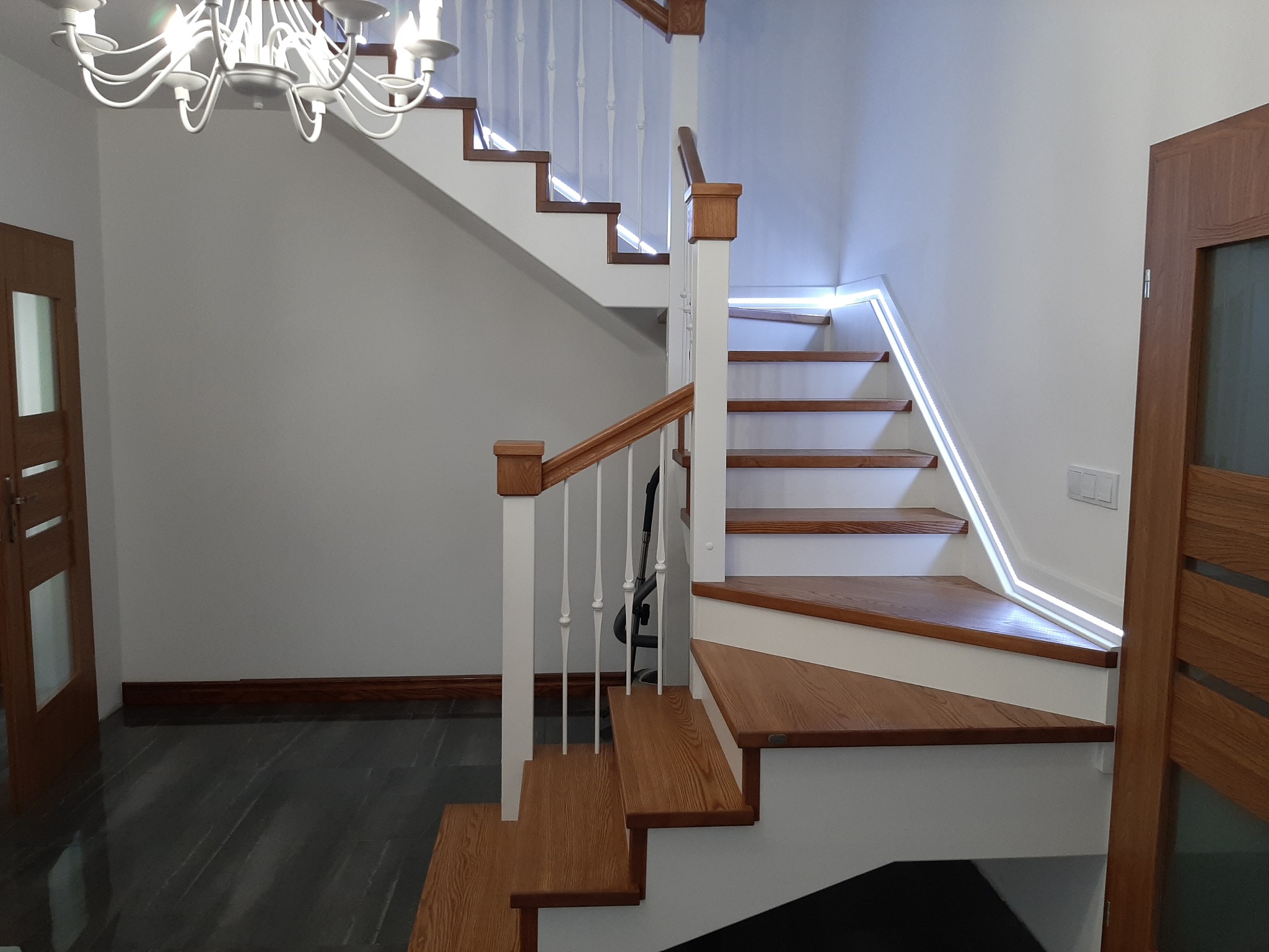 Dwubiegowe schody z drewnianymi stopniami i białymi podstopnicami, oświetlone taśmą LED, z białą balustradą i drewnianą poręczą, widoczne drzwi i żyrandol w tle.