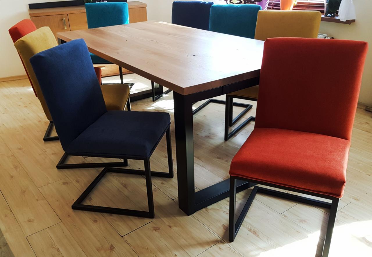 Stół z blatem z jasnego drewna i czarnymi, geometrycznymi nogami, otoczony krzesłami z tapicerką w odcieniach niebieskiego, czerwonego, żółtego i zielonego, na jasnej drewnianej podłodze.