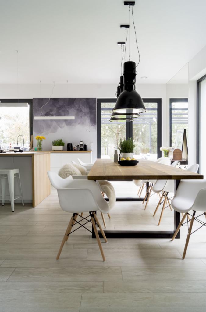 Jasna jadalnia z drewnianym stołem na metalowej podstawie, białymi krzesłami Eames z drewnianymi nogami i podłogą z paneli imitujących drewno, oświetlona trzema czarnymi lampami wiszącymi.