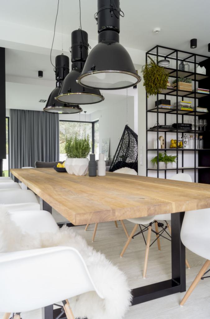 Stół z blatem z jasnego drewna na czarnych, metalowych nogach w nowoczesnej jadalni, nad nim wiszą trzy duże, czarne lampy przemysłowe, w tle regał z dekoracjami i fotel wiszący.