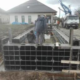 Wylewanie betonu do szalunków fundamentowych z użyciem pompy do betonu, w tle dom jednorodzinny w trakcie budowy.