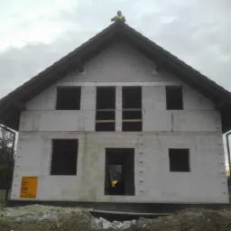 Surowy stan budynku z białych bloczków, widoczny pracownik na dachu w odblaskowej kamizelce i żółta tablica informacyjna na ścianie. Niezagospodarowany teren wokół.